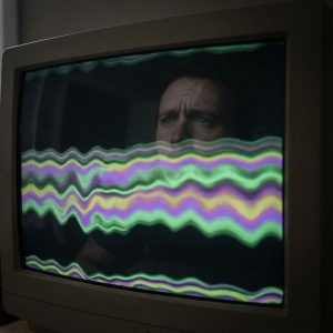 컴퓨터 화면에 일그러진 부자연스러운 색상 띠가 가득 차 있고, 그 위에 사람의 걱정스러운 얼굴이 희미하게 비친 모습이다.