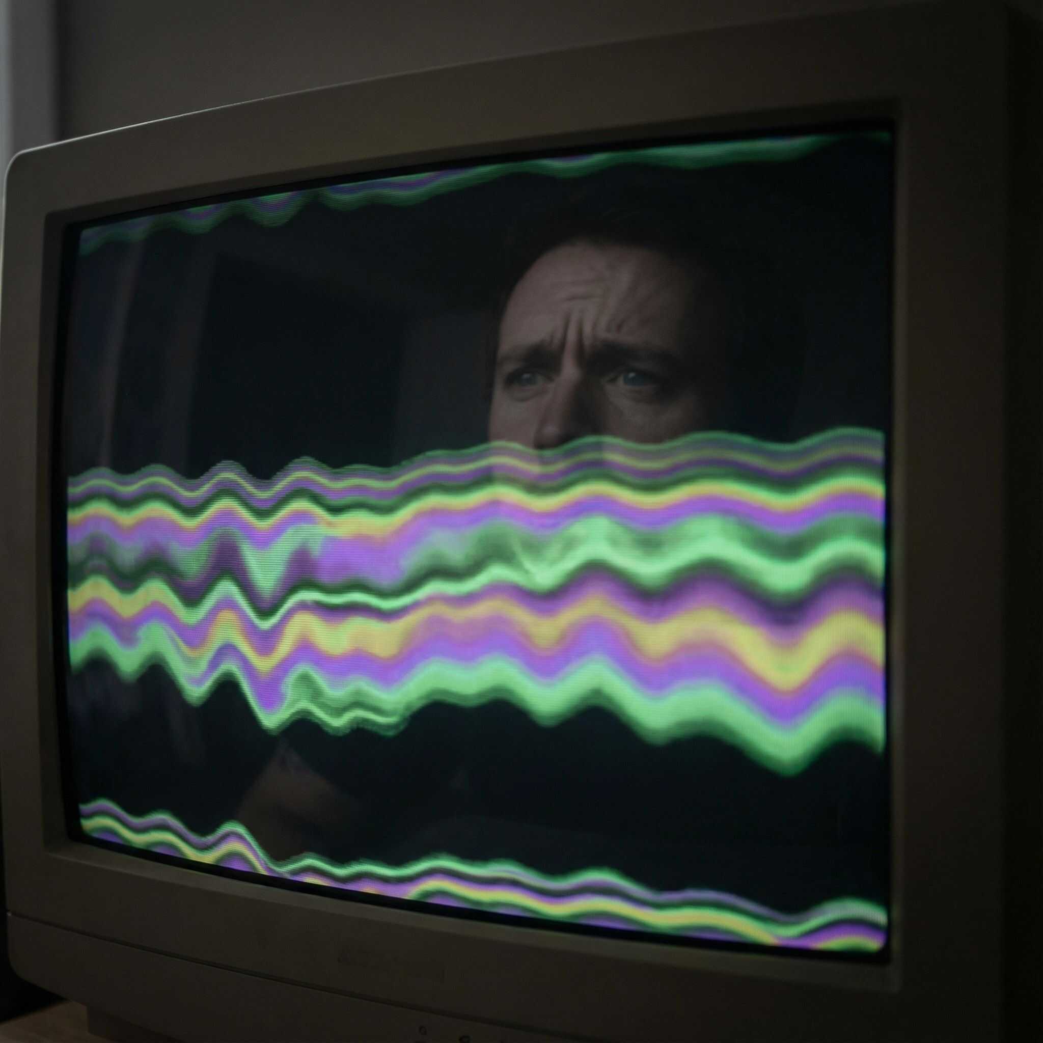 컴퓨터 화면에 일그러진 부자연스러운 색상 띠가 가득 차 있고, 그 위에 사람의 걱정스러운 얼굴이 희미하게 비친 모습이다.
